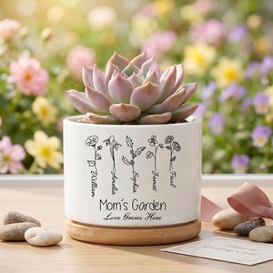 Puede incluir: Una maceta de cerámica blanca con una planta suculenta, sobre una base de madera. La maceta está decorada con dibujos lineales florales y el texto "Mom's Garden Love Grows Here" con nombres. Un lazo rosa y piedras decorativas están en primer plano.