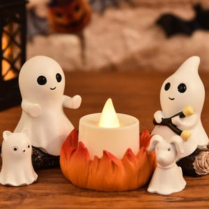 Peut inclure: Deux figurines de fantômes blancs aux yeux noirs, dont l'un tient un bâton, sont assises à côté d'une petite bougie chauffe-plat LED blanche placée dans un support circulaire orange et rouge en forme de flammes, avec un petit chien blanc.