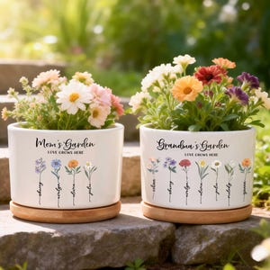 Pode incluir: Duas floreiras de cerâmica branca com pires de madeira, cada uma cheia de flores coloridas. Uma floreira diz "Mom's Garden" e a outra "Grandma's Garden". Ambas as floreiras têm desenhos florais e nomes.