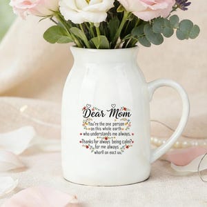 Op de afbeelding: Een witte keramische kan met een handvat, gevuld met een boeket van roze en witte rozen, lavendel en groen. De kan heeft de tekst "Dear Mom" en een liefdevolle boodschap. Verspreide bloemblaadjes en een parelketting staan op de voorgrond.