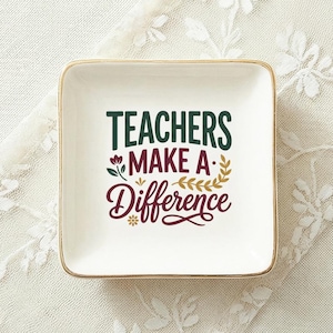 May include: A square white ceramic trinket dish with a gold-colored rim. The center features the text "TEACHERS MAKE A Difference" in dark green and maroon lettering, accented with small floral and leaf illustrations in green, maroon, and gold.