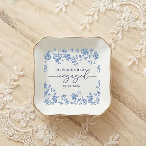 May include: A square, white ceramic ring dish with a gold rim, adorned with blue floral patterns. The dish features the text "OLIVIA & CHLOE engaged 05.30.2026". The dish is placed on a cream-colored lace cloth, with wedding rings and flowers nearby.