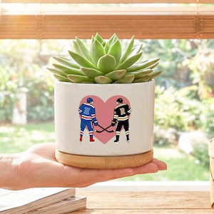 May include: A white ceramic planter with a succulent plant. The planter features a pink heart with hockey players in blue and black uniforms. The planter sits on a wooden base.