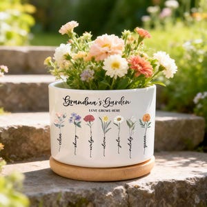 Könnte beinhalten: Ein weißer Keramik-Blumentopf mit dem Text "Grandma's Garden" und "Love Grows Here". Der Topf hat Blumenillustrationen und Namen. Er ist mit bunten Blumen gefüllt und steht im Freien auf einem Holzsockel.