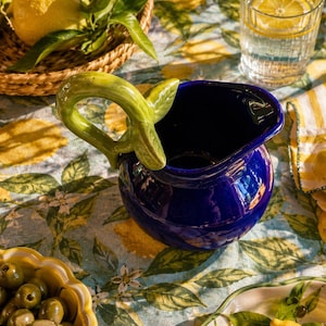 Puede incluir: Una jarra de cerámica azul intenso con un asa escultural en forma de rama con hojas verdes. La jarra tiene un cuerpo redondeado y un pico vertedor ancho, presentada como un recipiente funcional para servir líquidos en la mesa.
