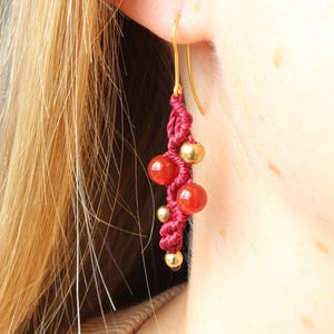Peut inclure: Boucle d'oreille artisanale avec un crochet doré, présentant un motif de cordon rouge tressé. La boucle d'oreille est ornée de perles rouges et dorées. La boucle d'oreille est portée à l'oreille d'une personne, mettant en valeur le design et la couleur du bijou.