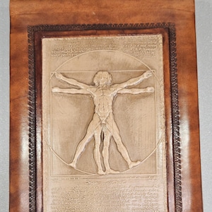 Pode incluir: Um caderno com capa de couro com um baixo-relevo do Homem Vitruviano de Leonardo da Vinci. A obra de arte é de cor bege clara, inserida numa moldura retangular com uma borda castanha mais escura. A capa do caderno é castanho médio.