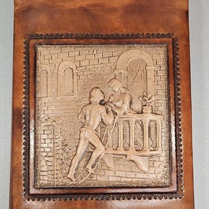 May include: A brown leather journal featuring an embossed scene of a man and woman on a balcony. The scene is framed by a decorative border and set against a brick wall background.