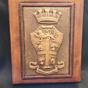 May include: A brown leather journal with a detailed embossed crest. The crest features a crown, shield, and banner with the inscription "NEI SECOLI FEDELE". The journal has a rectangular shape and a textured surface.