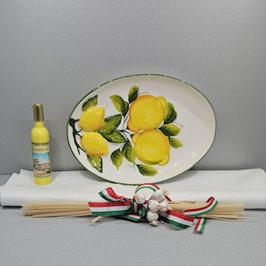 Puede incluir: Una fuente ovalada con un diseño de limón, una botella de Limoncello y un manojo de espaguetis atados con una cinta roja, blanca y verde. La fuente es blanca con un borde verde y los limones son amarillos.