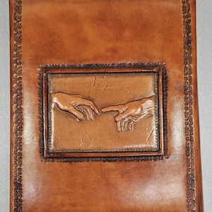 May include: A brown leather journal with a rectangular embossed design featuring two hands reaching towards each other. The journal has a decorative border and a classic, vintage aesthetic.