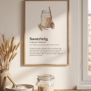Könnte beinhalten: Ein gerahmtes Wandbild mit einer wörterbuchartigen Definition von Sauerteig, inklusive einer Aquarellillustration eines Einmachglases mit Sauerteigstarter, einem Holzlöffel und Weizenähren auf einem hellen cremefarbenen Hintergrund.