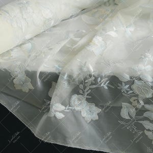 May include: Off-white fabric with delicate floral embroidery. The design features detailed roses and leaves in a soft, pale blue. The fabric is semi-sheer, with a rolled edge visible in the upper left corner. Ideal for bridal or formal wear.