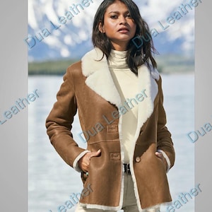 May include: A brown leather jacket featuring a cream-coloured shearling lining and collar. The jacket has two front pockets and a button closure. The model is wearing a cream-coloured turtleneck and light-coloured trousers. The background shows a mountain range and water.