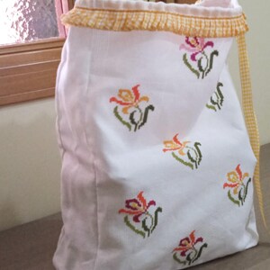 May include: A white fabric drawstring bag featuring a repeating cross-stitch pattern of multi-coloured flowers in shades of orange, pink, and green. The top opening is finished with a yellow and white gingham ruffled trim and a matching drawstring.
