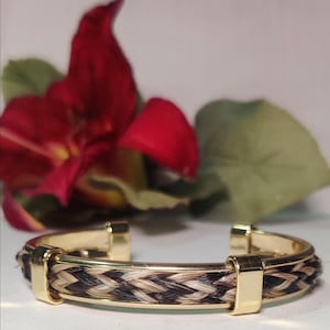 May include: A gold-toned metal cuff bracelet featuring a central braided inlay of dark brown, light brown, and cream-colored hair strands. The bracelet is secured with three polished gold-colored metal bands that hold the braided section in place.