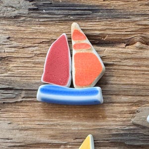 May include: A collection of colorful ceramic sailboat figurines arranged on a rustic wooden surface. The sailboats feature various designs and colors, including red, orange, yellow, pink, and blue. The sails are made from broken ceramic pieces.