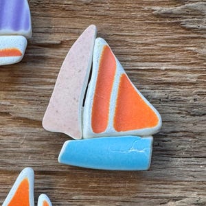 May include: A collection of colorful ceramic sailboat figurines with orange, blue, pink, and purple sails. The boats are arranged on a rustic wooden surface, with two small cloud-shaped accents.