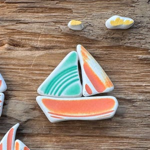 May include: A collection of handmade ceramic sailboat figurines in various colors, including red, orange, and green. Each boat features a unique striped sail design and sits on a white base. The sailboats are arranged on a weathered wooden surface.