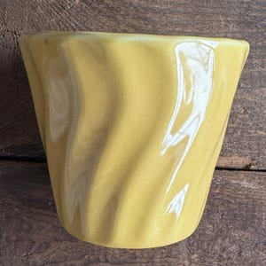 May include: A yellow ceramic planter with a wavy, textured design. The planter is empty and sitting on a wooden surface.