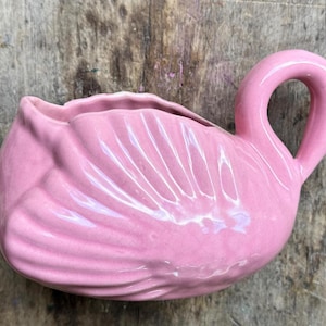 May include: A pink ceramic flamingo planter with a curved neck and open top. The planter has a textured surface with detailed feather impressions. The flamingo is set against a weathered wooden background.