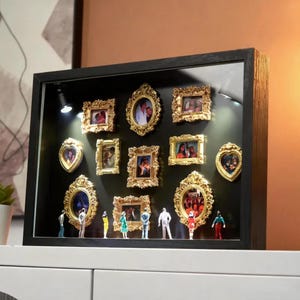 以下が含まれることがあります： 写真が入った小さな金色の装飾が施された額縁が複数ある黒いシャドーボックスディスプレイ。 ミニチュアのフィギュアがフレームの下に立っています。 ディスプレイは小さなスポットライトで照らされています。