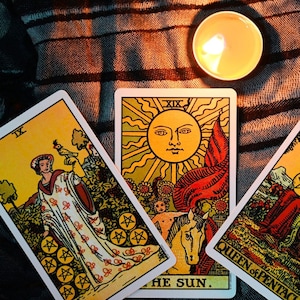 May include: A close-up of tarot cards with a lit candle. The cards feature illustrations of a woman, a sun with a face, and the words "THE SUN" and "QUEEN of PENTACLES". The cards are on a striped fabric.