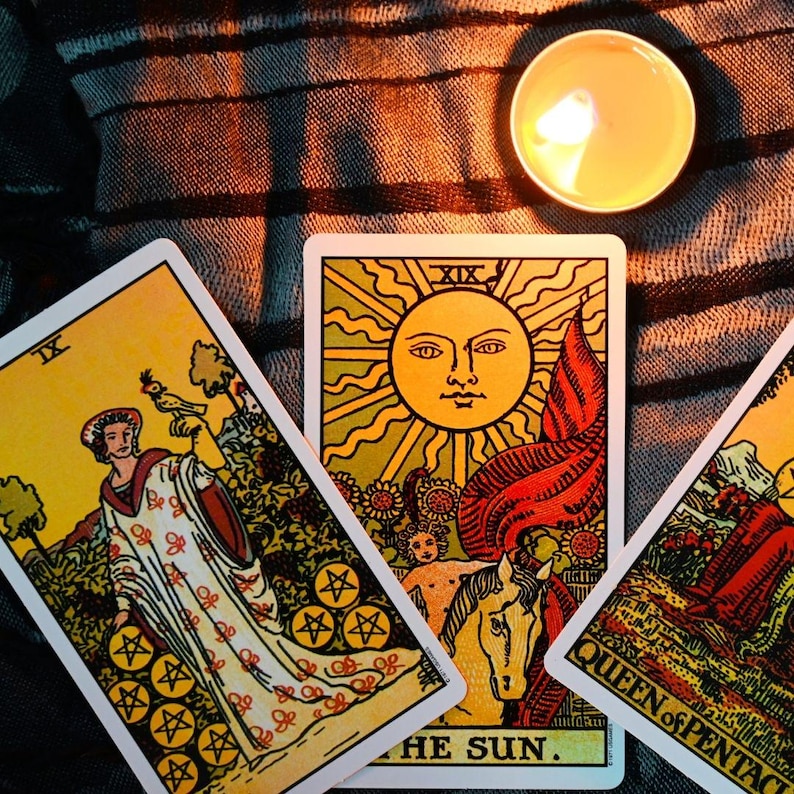 May include: A close-up of tarot cards with a lit candle. The cards feature illustrations of a woman, a sun with a face, and the words "THE SUN" and "QUEEN of PENTACLES". The cards are on a striped fabric.