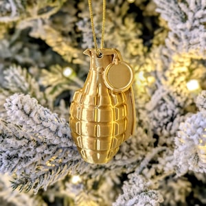 May include: A gold-colored Christmas ornament shaped like a hand grenade, hanging from a gold chain. The ornament has a textured surface and a small, round tag. The background is a blurred image of a snow-covered Christmas tree.