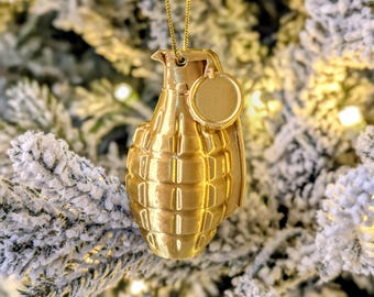 May include: A gold-colored Christmas ornament shaped like a hand grenade, hanging from a gold chain. The ornament has a textured surface and a small, round tag. The background is a blurred image of a snow-covered Christmas tree.