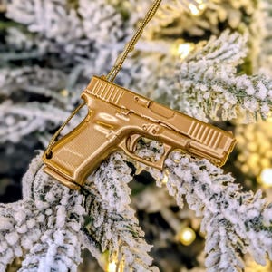 May include: A gold-colored handgun ornament hanging on a snow-covered Christmas tree branch. The ornament is a detailed replica of a firearm, with a textured grip and slide. The background is blurred, with hints of Christmas lights.