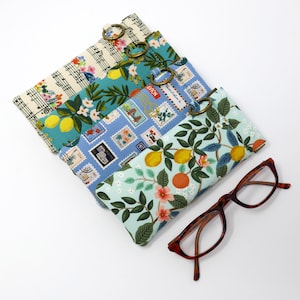 May include: Four different fabric eyeglass cases with floral and musical note patterns. The cases have a metal ring attached to the top. A pair of brown tortoiseshell eyeglasses are lying on a white surface.