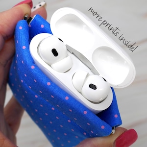 May include: A blue fabric case with pink polka dots, designed to hold a white wireless earbud case. The case is open, revealing the white earbuds inside. The text "more prints inside!" is written in black on a white background.