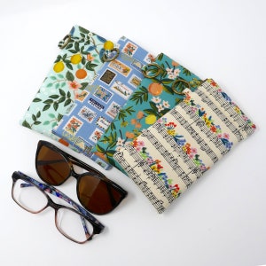 May include: Four different fabric eyeglass cases with floral and musical note patterns. The cases are stacked on top of each other. Two pairs of eyeglasses are also in the image.