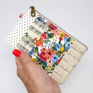 May include: A floral print zippered pouch with a patchwork design. The pouch features a mix of floral patterns, musical notes, and polka dots. The zipper is a bronze color.