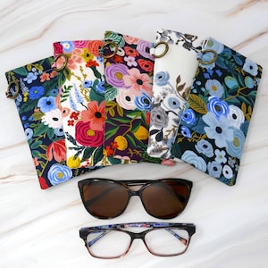 May include: Six colorful fabric eyeglass cases with floral patterns. The cases are lined up in a row, with the top case slightly overlapping the one below it. The cases are all closed, and the zipper pulls are visible. There are two pairs of eyeglasses in the image, one pair is brown and the other is black.