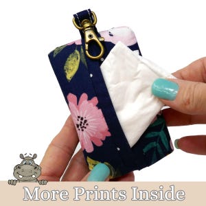 May include: A navy blue floral print wipe dispenser with a gold-colored clip. The dispenser features pink flowers and green leaves. White wipes are being pulled from the dispenser. The text "More Prints Inside" is at the bottom.