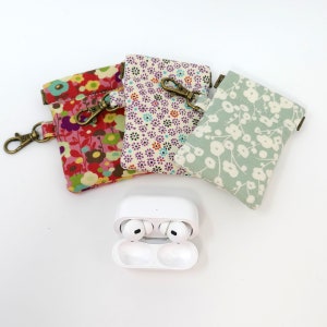 May include: Three small fabric pouches with floral patterns and a metal clasp. The pouches are arranged in a semi-circle with a white wireless charging case for earbuds in the center.