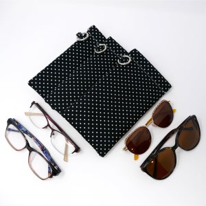 May include: A black and white polka dot fabric case with three silver rings for holding glasses. Two pairs of glasses are shown, one pair is brown and the other is clear.