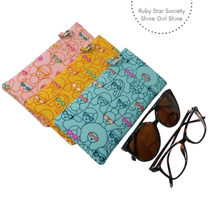 May include: Three rectangular fabric pouches with a repeating line art pattern of faces wearing sunglasses, in light pink, yellow, and teal. Each pouch features a metal split ring. Text reads Ruby Star Society Shine Girl Shine. Two pairs of glasses.