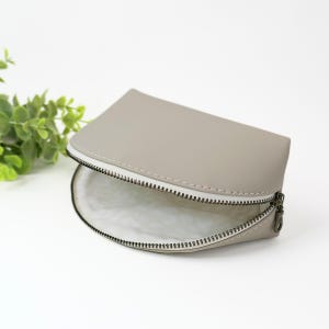 May include: A semi-circular pouch in a solid light taupe color with a smooth exterior finish. The pouch features a white zipper closure with a dark metal pull tab and a white interior lining that has a subtle, repeating textured pattern.