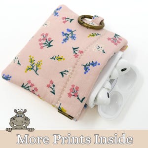 May include: A pink floral print pouch with a bronze-colored clasp and ring, holding a white wireless earbud case. The pouch is decorated with small flowers in blue, yellow, and pink. The text "More Prints Inside" is at the bottom.