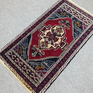 Op de afbeelding: Een rechthoekig, handgeweven tapijt met een centraal rood medaillonontwerp. Het tapijt heeft ingewikkelde patronen in marineblauw, crème en bordeaux, met een crèmekleurige franje langs de randen. Het tapijt ligt op een lichtgrijze ondergrond.