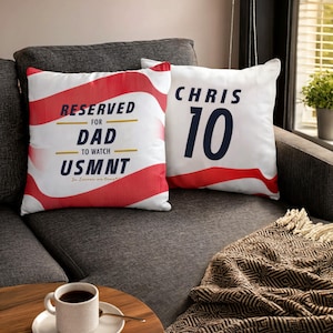 Puede incluir: Dos cojines cuadrados de tela blanca con rayas rojas onduladas. Uno presenta el texto RESERVED FOR DAD TO WATCH USMNT In Soccer we trust. El otro muestra el nombre CHRIS y el número 10 en una fuente grande de color azul oscuro.