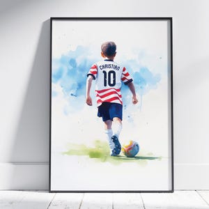 May include: Watercolour print of a soccer player in a red and white striped jersey with the name "Christian" and the number 10 on the back. The player is kicking a football on a green field, with blue watercolour accents in the background.
