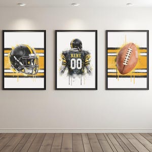 May include: Set of three framed art prints featuring watercolor illustrations of football-themed elements. Includes a black and gold helmet, a football player's jersey with the number 00, and a brown football. Each print has a white and yellow striped background.