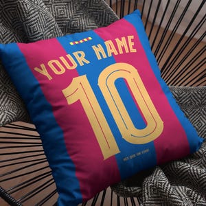 Op de afbeelding: Een blauw en roze kussen met een geel nummer 10 en de tekst "YOUR NAME" in geel. Het kussen heeft een blauw en roze gestreept patroon en de tekst "MES QUE UN CLUB" in wit.