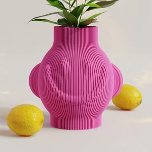 Könnte beinhalten: Eine rosa Keramikvase mit einem lächelnden Gesichtsdesign. Die Vase hat eine gerippte Textur und ist mit zwei kleinen Ohren auf jeder Seite verziert. Die Vase ist mit grünen Pflanzenblättern gefüllt und steht auf einer weißen Oberfläche mit zwei Zitronen.