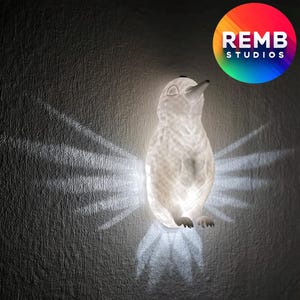 Puede incluir: Una luz nocturna de pingüino de cerámica blanca con una superficie texturizada. La luz está encendida y proyecta un brillo cálido en la pared detrás de ella.