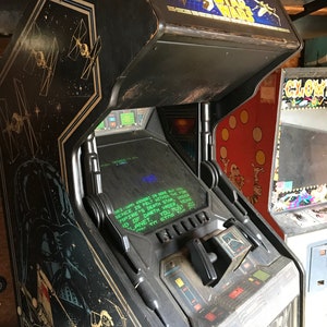 May include: A vintage Star Wars arcade game with a black cabinet and a screen displaying the text "JBT-LAN KENDORS DEBUT M SENCE IS FELT WITHIN THE FOR MPTRE'S DEATH STAR UNDER 'D OF DARTH VADER, NEAR LANET. YOU MUST JOI".